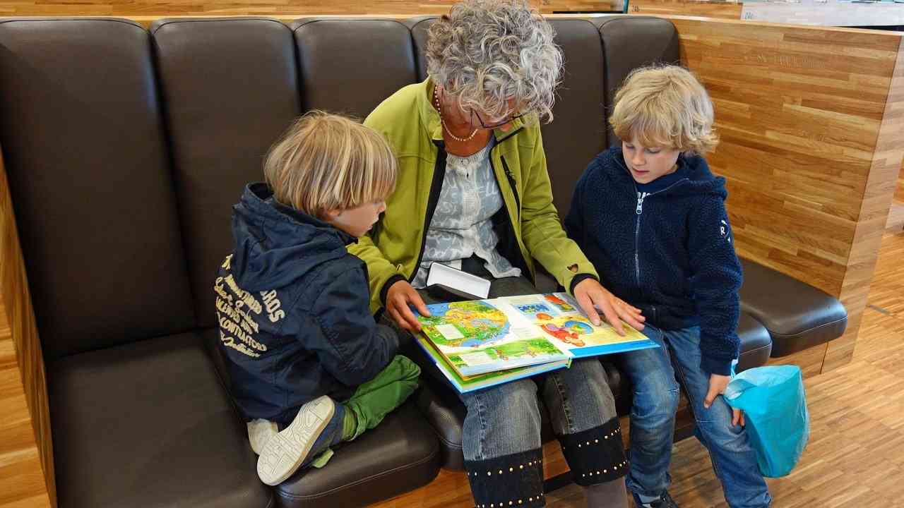 Бабушка с внуком и внучкой сидит на диване и читает вслух книгу